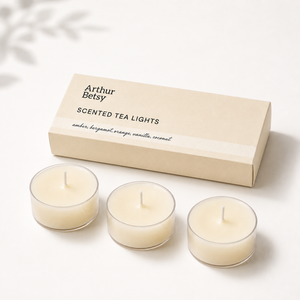 Three cream-colored scented tea lights in clear glass holders are arranged in front of their beige packaging box, which is printed with the brand name 'Arthur Betsy' and the scent description 'Scented Tea Lights'.