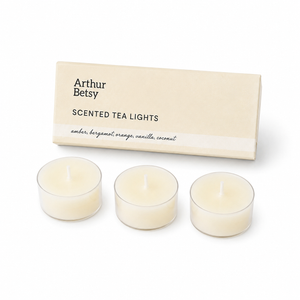 Three small tea light candles in clear holders with cream-colored wax, displayed in front of a beige Arthur Betsy box labeled 'Scented Tea Lights' on a white background. Tropical Breeze scented tea lights with amber, bergamot, orange, coconut and vanilla fragrance notes for a warm, relaxing ambience.