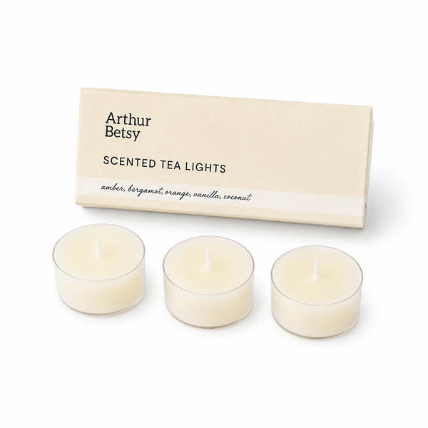 Three small tea light candles in clear holders with cream-colored wax, displayed in front of a beige Arthur Betsy box labeled 'Scented Tea Lights' on a white background. Tropical Breeze scented tea lights with amber, bergamot, orange, coconut and vanilla fragrance notes for a warm, relaxing ambience.