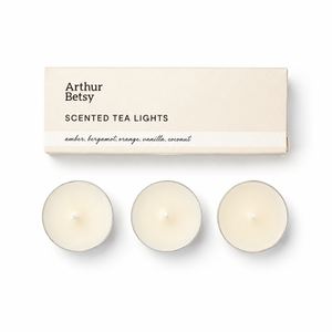 Three small tea light candles displayed below their cream-colored packaging box, which is labeled 'Arthur Betsy SCENTED TEA LIGHTS'. Arthur Betsy set of three scented tea lights made with a premium soy and mineral wax blend, presented in a slim gift-ready box.