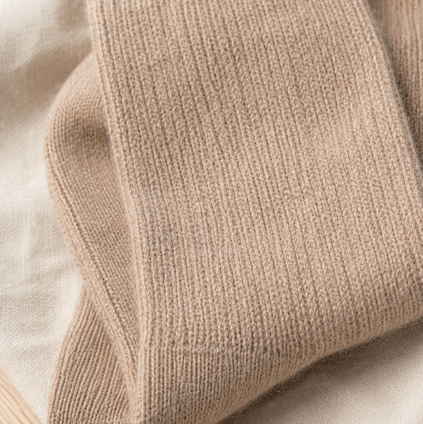 A close-up of folded ribbed knit socks in a warm beige color, resting on a soft cream fabric background. Soft neutral taupe wool socks made from alpaca wool, nylon and spandex—a luxurious winter accessory and thoughtful self-care gift.