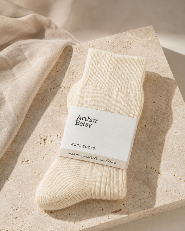 A single cream-colored wool sock wrapped in a white paper band labeled 'Arthur Betsy', styled on a beige stone slab with draped linen fabric. Soft cream socks made with wool, acrylic and spandex, combining warmth, stretch and lasting comfort.