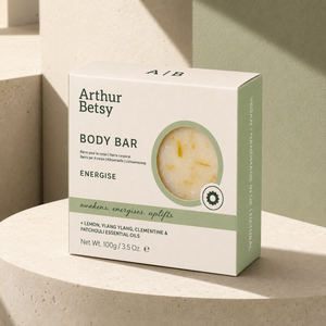Arthur Betsy Energise body bar box in a light cream and sage green color, displayed on a textured beige stone pedestal with soft shadows. Staffordshire-made natural soap bar with refreshing citrus and botanical notes, presented in minimal recyclable packaging and ideal for self-care gifting.
