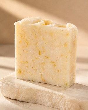 A rectangular bar of handmade soap with a creamy off-white color and small yellow botanical flecks, displayed on a light stone slab against a warm beige background. Natural citrus soap bar with nourishing olive oil, coconut oil, shea butter, castor oil and sweet almond oil for gentle everyday cleansing.