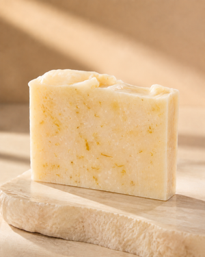 A rectangular bar of handmade soap in a pale creamy yellow color with small golden-yellow botanical inclusions, displayed on a rustic beige stone slab against a warm beige background with soft diagonal shadows. Natural citrus soap bar with nourishing olive oil, coconut oil, shea butter, castor oil and sweet almond oil for gentle everyday cleansing.