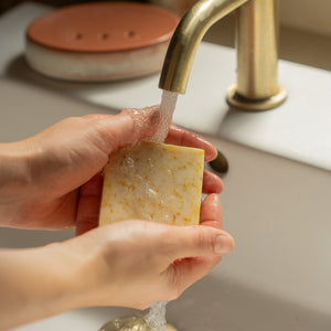 Person holding a bar of soap under running water from a gold faucet. Handcrafted lemon and clementine body soap with floral ylang ylang, earthy patchouli and calendula flowers for an uplifting aromatic cleanse.