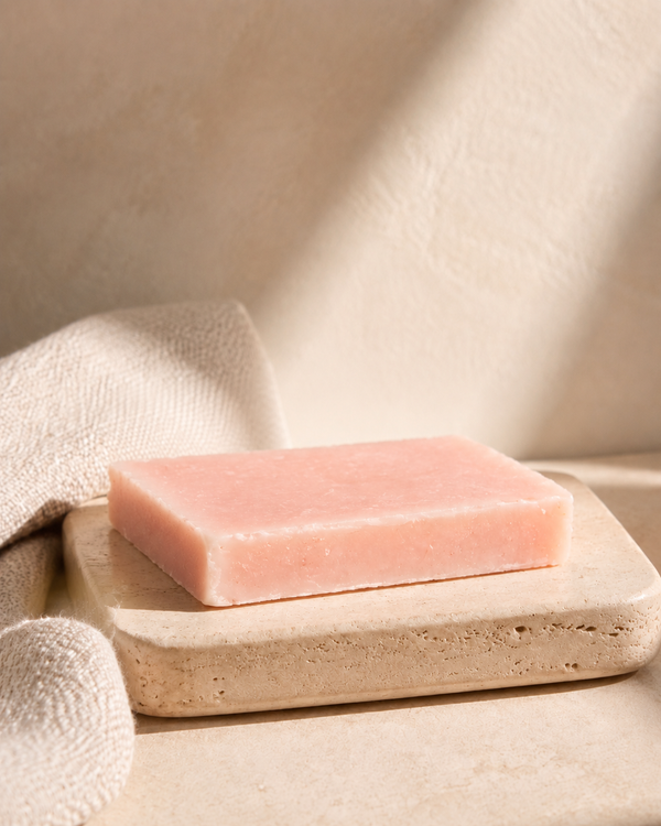 A rectangular pink soap bar resting on a beige stone dish, with a soft beige towel draped nearby in warm natural light. Natural cold-process soap bar with nourishing olive oil, coconut oil, shea butter, castor oil and sweet almond oil for gentle everyday cleansing.