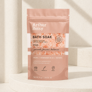 Arthur Betsy Focus Bath Soak in a stand-up pouch, displayed on a white pedestal against a warm beige background with soft shadows. Calming single-bath salt sachet with fresh eucalyptus and warm woody notes, perfect for travelling, self-care or wellbeing gifting.