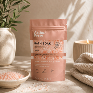 A stand-up pouch of Arthur Betsy Focus Bath Soak stands on a beige surface in a styled spa-like setting with a lit candle, a bowl of pink bath salts, and a plant in the background. Handmade herbal bath salts with mineral-rich salts and pure essential oils, presented in a printed single-use bath soak pouch.