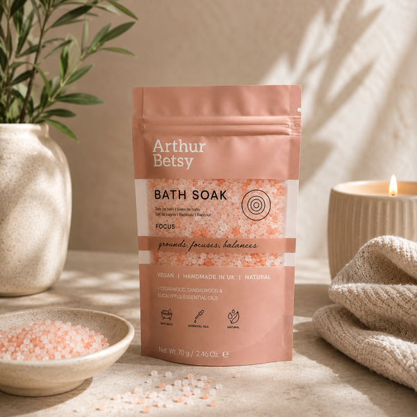 A stand-up pouch of Arthur Betsy Focus Bath Soak stands on a beige surface in a styled spa-like setting with a lit candle, a bowl of pink bath salts, and a plant in the background. Handmade herbal bath salts with mineral-rich salts and pure essential oils, presented in a printed single-use bath soak pouch.