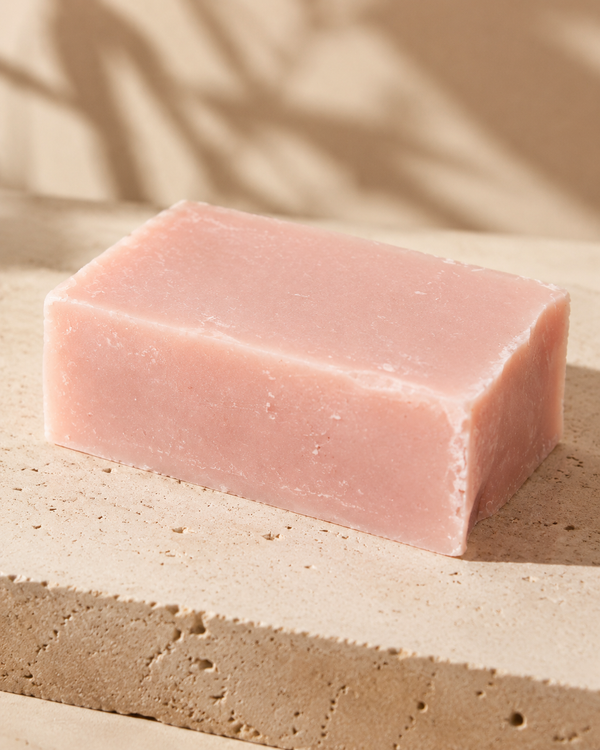 A rectangular bar of pale pink soap resting on a textured beige stone slab with soft shadow overlays. Natural woody soap bar with nourishing olive oil, coconut oil, shea butter, castor oil and sweet almond oil for gentle everyday cleansing.
