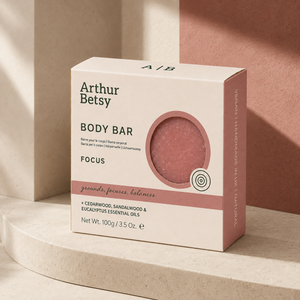 Arthur Betsy Focus Body Bar in a cream-colored box with dusty rose accents, displayed on a curved beige stone pedestal against a terracotta background. Artisan body soap made with plant-based oils, naturally retained glycerin, madder root powder and pure essential oils to leave skin feeling cleansed and refreshed.