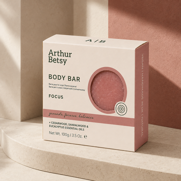 Arthur Betsy Focus Body Bar in a cream-colored box with dusty rose accents, displayed on a curved beige stone pedestal against a terracotta background. Artisan body soap made with plant-based oils, naturally retained glycerin, madder root powder and pure essential oils to leave skin feeling cleansed and refreshed.