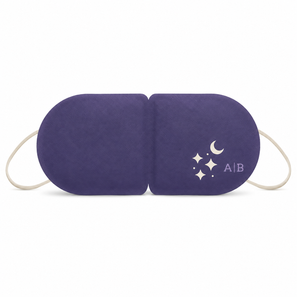 A purple sleep mask with white elastic ear loops and a white crescent moon and star graphic on the right side, shown on a plain white background. Unscented warming eye mask that activates when opened, offering calming heat for relaxation, bedtime, travel or flights.