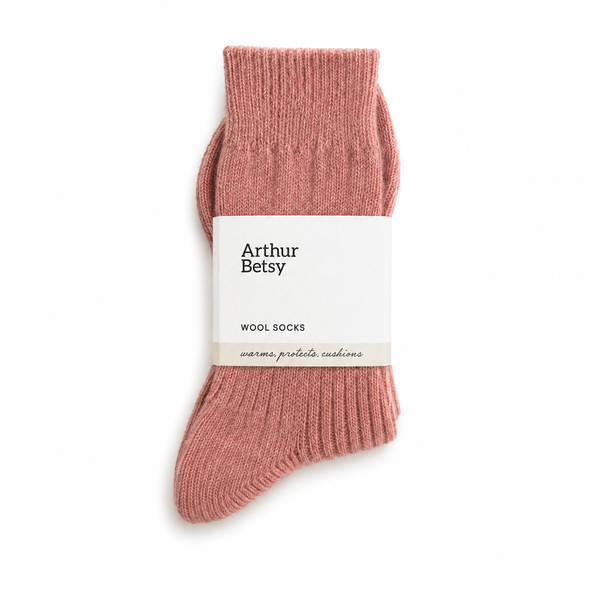 A pair of ribbed knit wool socks in a dusty rose pink color, wrapped in a white paper band with the brand name 'Arthur Betsy' printed on it, set against a plain white background. Arthur Betsy luxury alpaca wool socks in muted rose, made with an 80% alpaca wool blend for warmth, softness and everyday comfort.