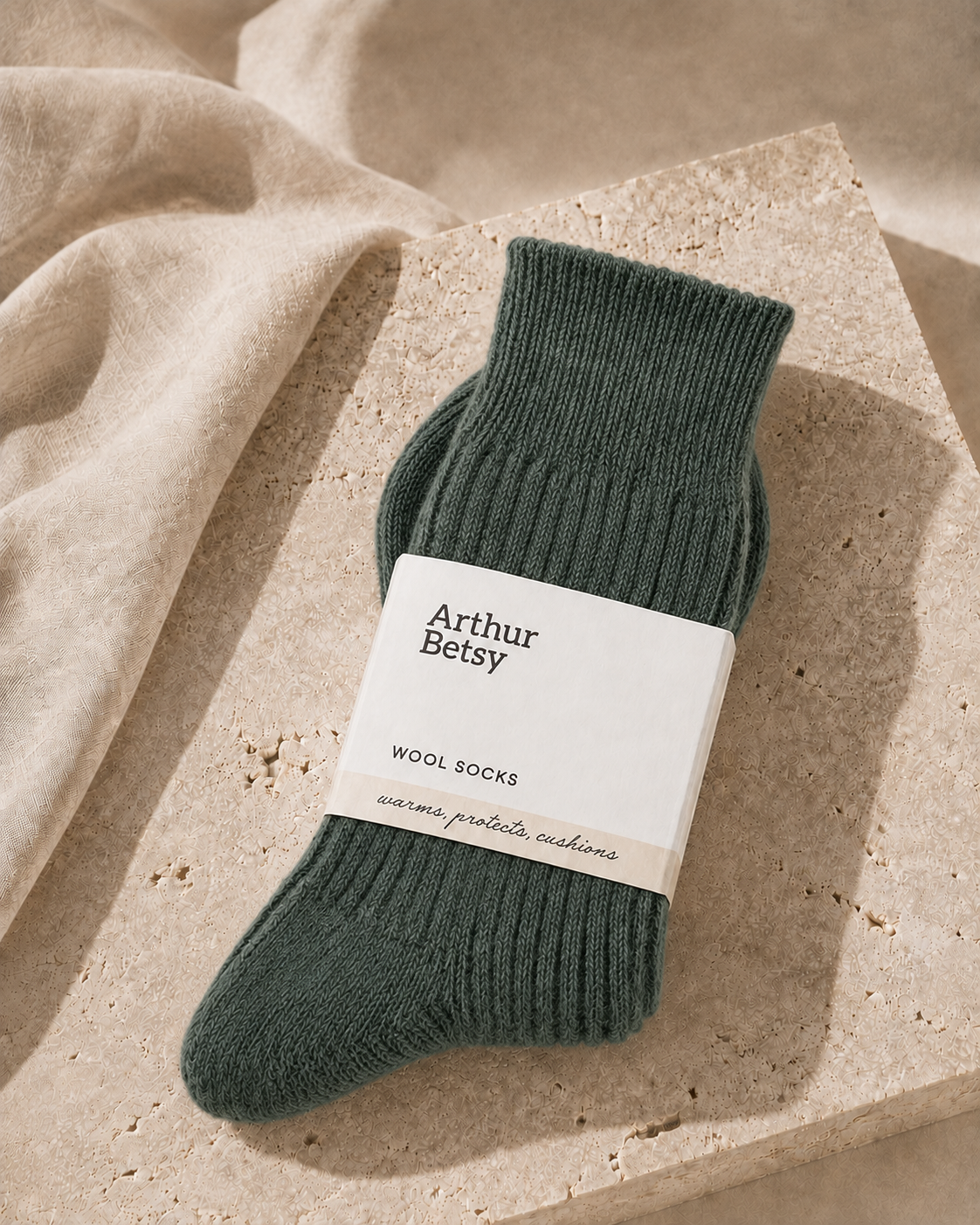 A pair of ribbed wool socks in a muted dark green, wrapped with a white paper label reading 'Arthur Betsy', styled on a textured stone slab with a beige linen cloth. Cosy Arthur Betsy alpaca socks in Nordic Pine, ideal as a birthday, Christmas, stocking filler or wellbeing gift.