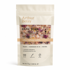 A stand-up pouch of Arthur Betsy Bath Soak in the Restore scent, featuring a cream-colored package with a clear window showing pink and purple bath salts inside. Calming single-bath salt sachet with soothing lavender, herbal rosemary and warming cardamom notes, perfect for travelling, pampering or wellbeing gifting.
