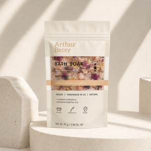 A stand-up pouch of Arthur Betsy Bath Soak placed on a light stone pedestal in a sunlit setting. The package is off-white with tan text and a central photo window showing pink and purple bath salts. Handmade herbal bath salts with dried calendula, lavender flowers and rose petals, presented in a printed single-use bath soak pouch.