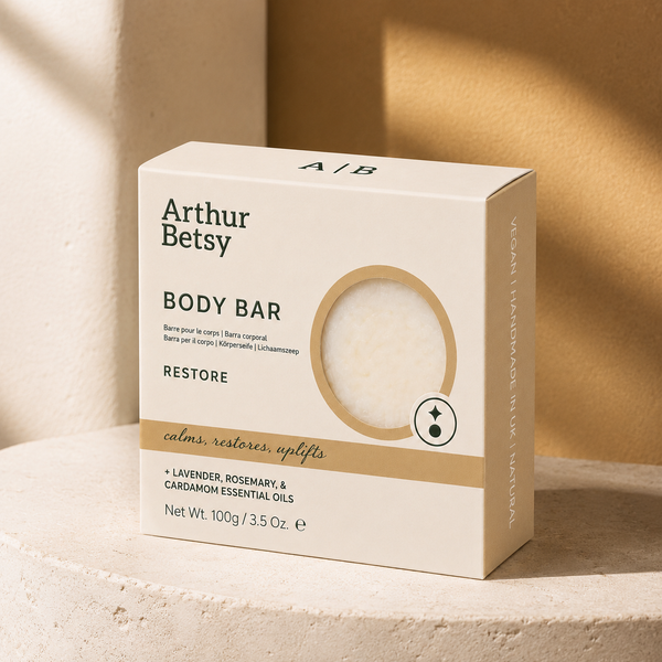 Arthur Betsy Restore Body Bar box in a warm cream color, featuring a circular cutout window that reveals the white bar inside. The box is styled on a textured stone pedestal with warm, sunlit shadows. Staffordshire-made lavender, rosemary and cardamom soap in minimal recyclable packaging—a thoughtful birthday, Christmas or self-care gift.