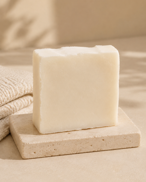 A rectangular bar of creamy off-white soap standing upright on a light stone dish, with a folded beige towel in the background and soft shadow lighting. Artisan body soap made with plant-based oils, naturally retained glycerin and pure essential oils to leave skin feeling soft and refreshed.