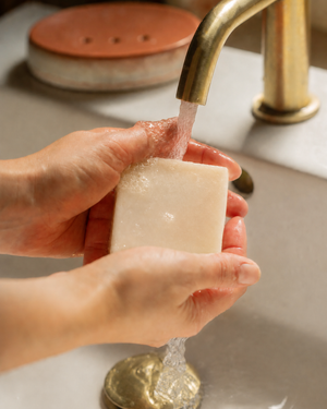 A close-up of hands washing a rectangular bar of soap under running water from a brass faucet in a sink. Handcrafted cold-process soap with calming lavender, refreshing rosemary and warm cardamom for a soothing, aromatic bathing ritual.
