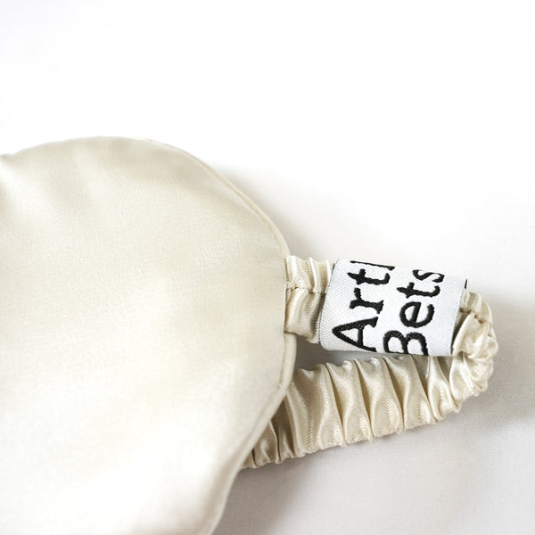 Close-up of a cream-colored satin sleep mask with a gathered elastic strap and a white fabric label that reads 'Arthur Betsy'. Soft and lightweight satin eye mask for women, a thoughtful self-care, birthday, Christmas or wellbeing gift for better rest.