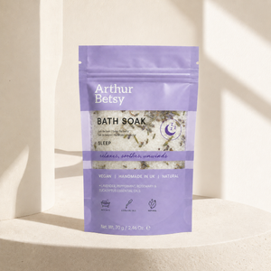 A stand-up pouch of Arthur Betsy Bath Soak in the 'Sleep' scent, featuring a lavender purple design with white text and a clear window showing dried botanicals inside, set against a warm beige background with soft diagonal shadows. Single-bath Arthur Betsy sleep soak in a travel-friendly sachet—a thoughtful relaxation, pampering or wellbeing gift for restful evenings.