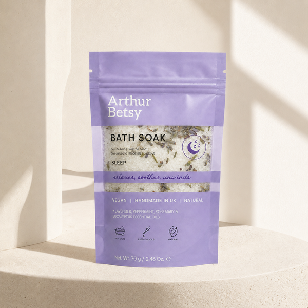 A stand-up pouch of Arthur Betsy Bath Soak in the 'Sleep' scent, featuring a lavender purple design with white text and a clear window showing dried botanicals inside, set against a warm beige background with soft diagonal shadows. Single-bath Arthur Betsy sleep soak in a travel-friendly sachet—a thoughtful relaxation, pampering or wellbeing gift for restful evenings.