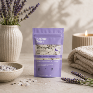 Arthur Betsy Sleep Calming Lavender Bath Soak in a lavender pouch, styled on a neutral stone surface with lavender sprigs, a bowl of bath salts, a candle, and a folded towel. Herbal sleep bath salts with Epsom salts and dried lavender, ideal for soothing tired muscles, easing tension and helping the body unwind before bedtime.