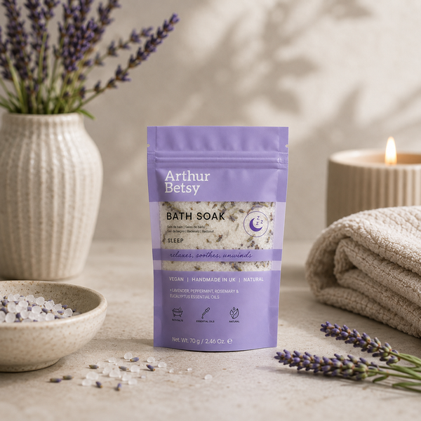 Arthur Betsy Sleep Calming Lavender Bath Soak in a lavender pouch, styled on a neutral stone surface with lavender sprigs, a bowl of bath salts, a candle, and a folded towel. Herbal sleep bath salts with Epsom salts and dried lavender, ideal for soothing tired muscles, easing tension and helping the body unwind before bedtime.