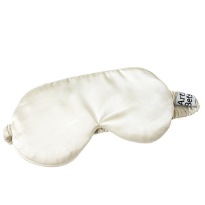 An off-white satin sleep mask with elasticized ear loops and a small black and white fabric tag on the side, shown on a solid white background. Arthur Betsy satin sleeping mask made from 100% polyester—ideal for beauty sleep, flights, holidays and peaceful nights at home.