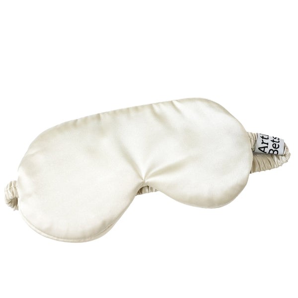 An off-white satin sleep mask with elasticized ear loops and a small black and white fabric tag on the side, shown on a solid white background. Arthur Betsy satin sleeping mask made from 100% polyester—ideal for beauty sleep, flights, holidays and peaceful nights at home.