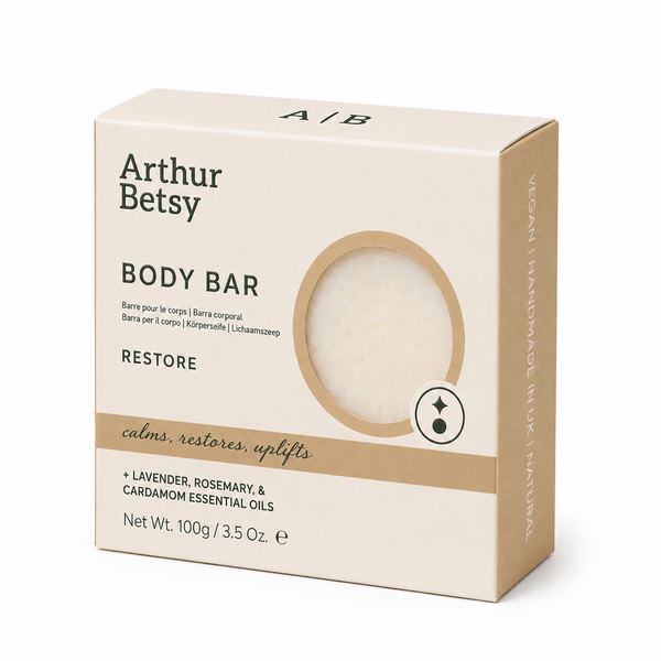Arthur Betsy Restore Body Bar packaging box in a warm beige color, featuring a circular cutout window that reveals the white bar of soap inside. The box includes text for lavender, rosemary, and cardamom essential oils. Arthur Betsy 100g handmade body soap with lavender, rosemary and cardamom essential oils, hand-poured in Staffordshire.