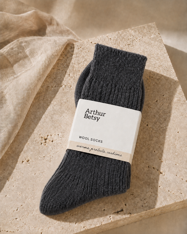 A pair of dark charcoal gray ribbed wool socks wrapped in a white paper band labeled 'Arthur Betsy', styled on a textured beige stone slab with draped linen fabric. Cosy Arthur Betsy alpaca socks in Twilight, ideal as a birthday, Christmas, stocking filler or wellbeing gift.