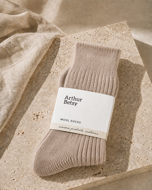A single ribbed knit sock in a warm taupe-beige color, wrapped in a white paper band labeled 'Arthur Betsy WOOL SOCKS', styled on a textured stone slab with a linen cloth in the background. Cosy Arthur Betsy alpaca socks in Wheat Taupe, ideal as a birthday, Christmas, stocking filler or wellbeing gift.