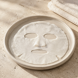 A white sheet mask with cutouts for the eyes, nose, and mouth, resting on a round beige ceramic plate on a textured beige stone surface with a folded towel in the background. Hydrating face sheet mask for all skin types, formulated to nourish the skin and reduce the appearance of fine lines for a smoother, radiant complexion.