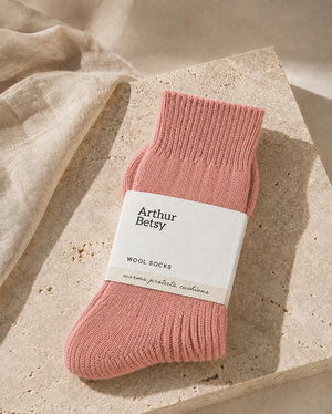A single pink knit sock with a white paper band reading 'Arthur Betsy WOOL SOCKS' is styled on a beige stone slab with soft linen fabric in the background. Cosy Arthur Betsy alpaca socks in muted rose, ideal as a birthday, Christmas, stocking filler or wellbeing gift for her.