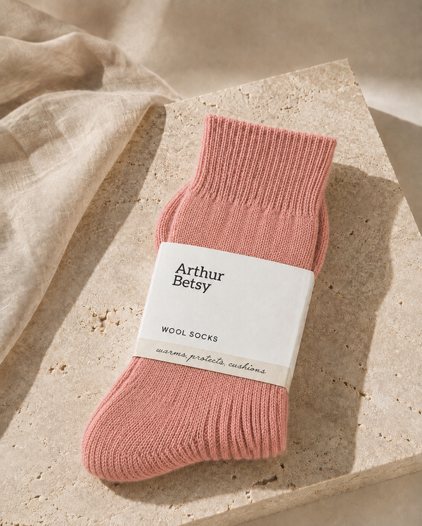 A single pink knit sock with a white paper band reading 'Arthur Betsy WOOL SOCKS' is styled on a beige stone slab with soft linen fabric in the background. Cosy Arthur Betsy alpaca socks in muted rose, ideal as a birthday, Christmas, stocking filler or wellbeing gift for her.