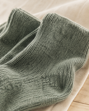 A close-up of a ribbed knit sock in a muted sage green color, resting on a soft beige fabric surface. Nordic Pine alpaca wool-blend socks in a comfortable one-size fit, designed for cosy lounging and stylish everyday wear.
