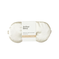 An off-white satin sleep eye mask by Arthur Betsy, wrapped in a white paper band with black text and a beige accent stripe, set against a plain white background. Arthur Betsy luxury satin eye mask designed to block out light and support restful, uninterrupted sleep at home or while travelling.