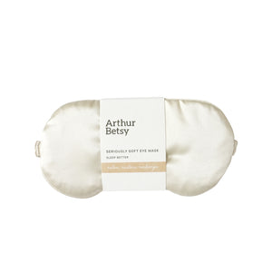 An off-white satin sleep eye mask by Arthur Betsy, wrapped in a white paper band with black text and a beige accent stripe, set against a plain white background. Arthur Betsy luxury satin eye mask designed to block out light and support restful, uninterrupted sleep at home or while travelling.