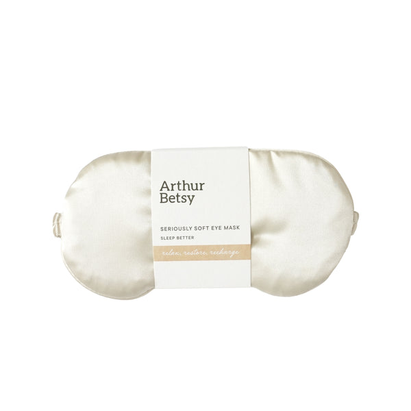 An off-white satin sleep eye mask by Arthur Betsy, wrapped in a white paper band with black text and a beige accent stripe, set against a plain white background. Arthur Betsy luxury satin eye mask designed to block out light and support restful, uninterrupted sleep at home or while travelling.