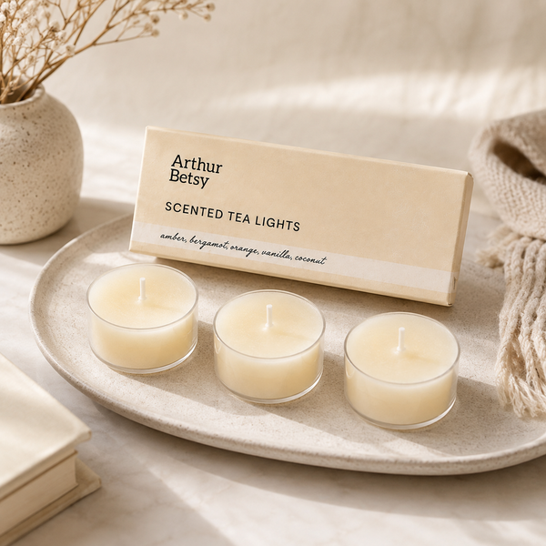 Three small tea light candles in clear glass holders sit on a round tray beside their cream-colored Arthur Betsy box labeled 'Scented Tea Lights'. The scene is styled with a woven throw, a vase of dried stems, and a book on a soft neutral fabric background. Boxed set of three luxury scented tea-light candles—an ideal self-care treat, stocking filler, Christmas gift or addition to a wellbeing hamper.