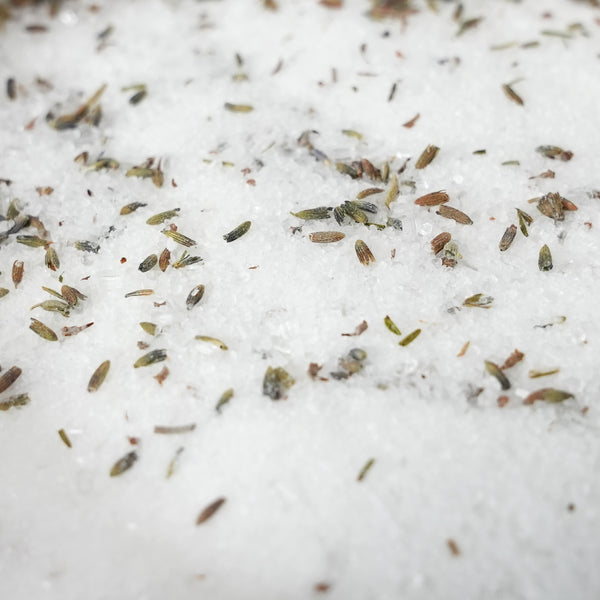 Close-up of coarse white salt crystals scattered with small dried green and brown herb seeds on top. Relaxing aromatherapy bath soak with lavender, peppermint, eucalyptus and rosemary essential oils, designed for a calming evening self-care ritual.