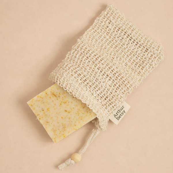 A cream-colored cotton mesh soap bag with a drawstring and wooden bead, containing a speckled yellow soap bar, photographed on a solid beige background. Natural sisal soap saver bag designed to improve lather, provide gentle body exfoliation and help make the most of every soap bar.