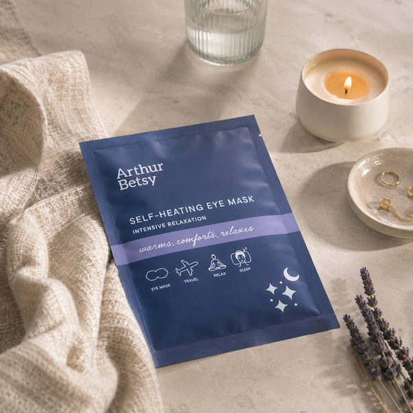 Arthur Betsy Self-Heating Eye Mask in a blue and purple foil packet, styled on a light stone surface with a beige linen cloth, a lit candle, a glass, and lavender sprigs. Soft disposable warming eye mask for relaxation and sleep, ideal for unwinding before bed, relieving tired eyes or enjoying an on-the-go moment of calm. warming eye masks