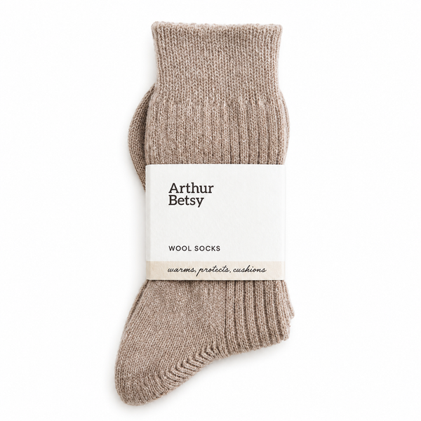 A pair of ribbed knit wool socks in a warm taupe-beige color, wrapped in a white paper band with the brand name 'Arthur Betsy' printed on it, set against a plain white background. Arthur Betsy luxury alpaca wool socks in Wheat Taupe, made with an 80% alpaca wool blend for warmth, softness and everyday comfort.